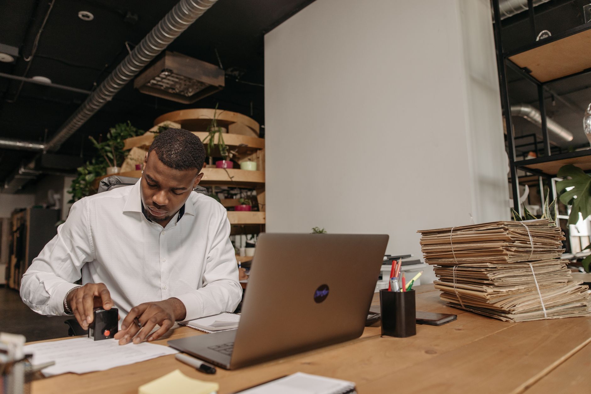 Homme afro-américain concentré sur l'apposition de tampons sur des documents à un bureau avec un ordinateur portable et des dossiers.