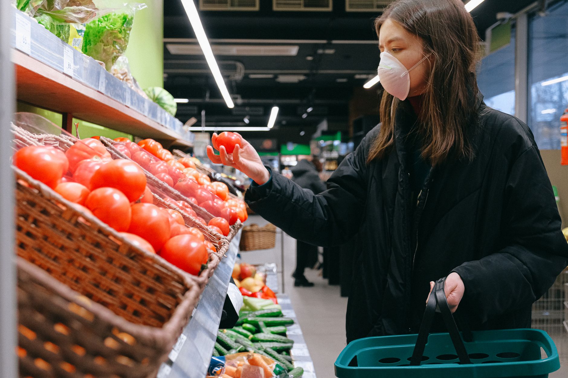 Une femme portant un masque sélectionne des tomates dans un magasin d'alimentation, illustrant les habitudes d'achat pendant la pandémie.