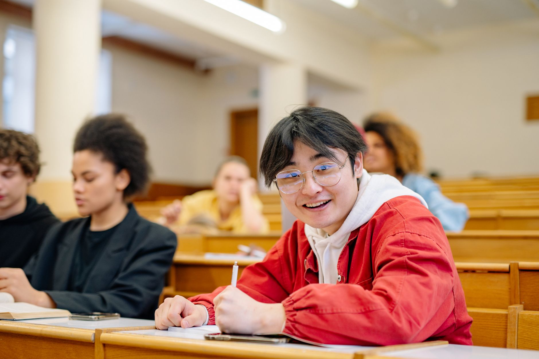 Étudiant souriant en veste rouge, stylo à la main, assis à un pupitre en bois dans un amphithéâtre, avec d’autres étudiants flous autour.