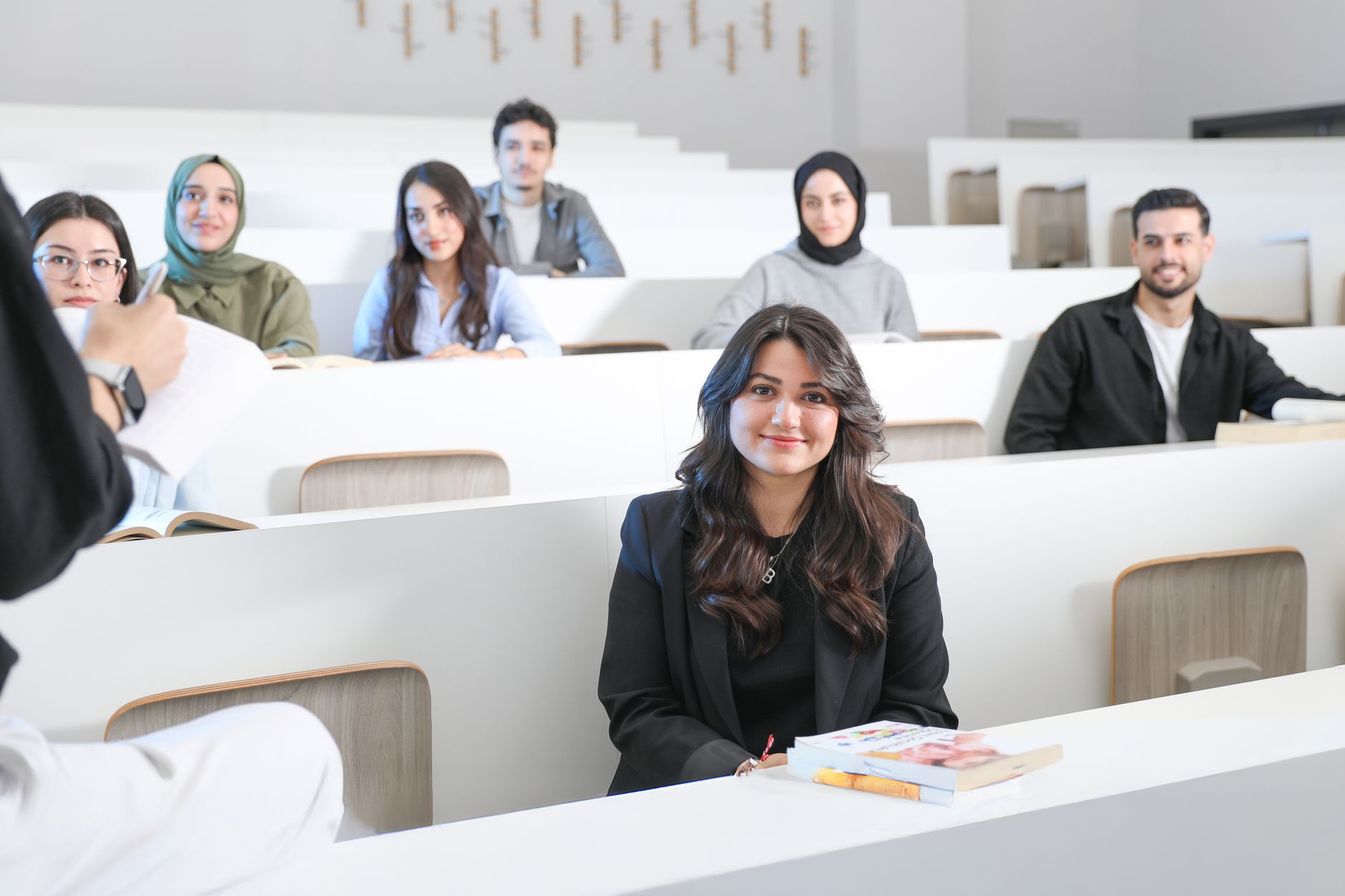 Étudiants assis dans un amphithéâtre aux bancs blancs, une jeune femme souriante au premier plan avec des livres posés devant elle.