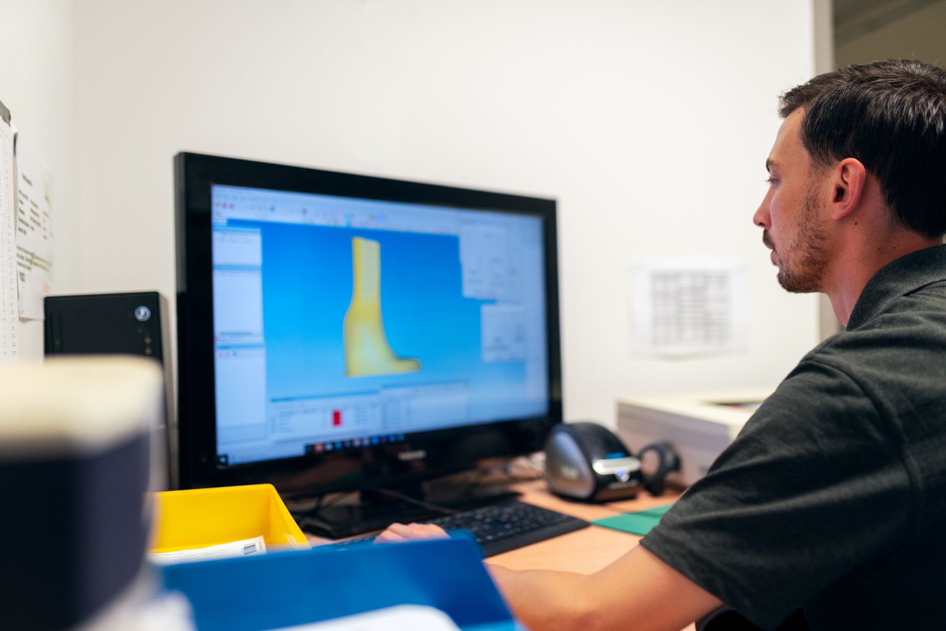 Homme de profil devant un écran d’ordinateur affichant un modèle 3D jaune dans un logiciel de conception, sur un bureau.