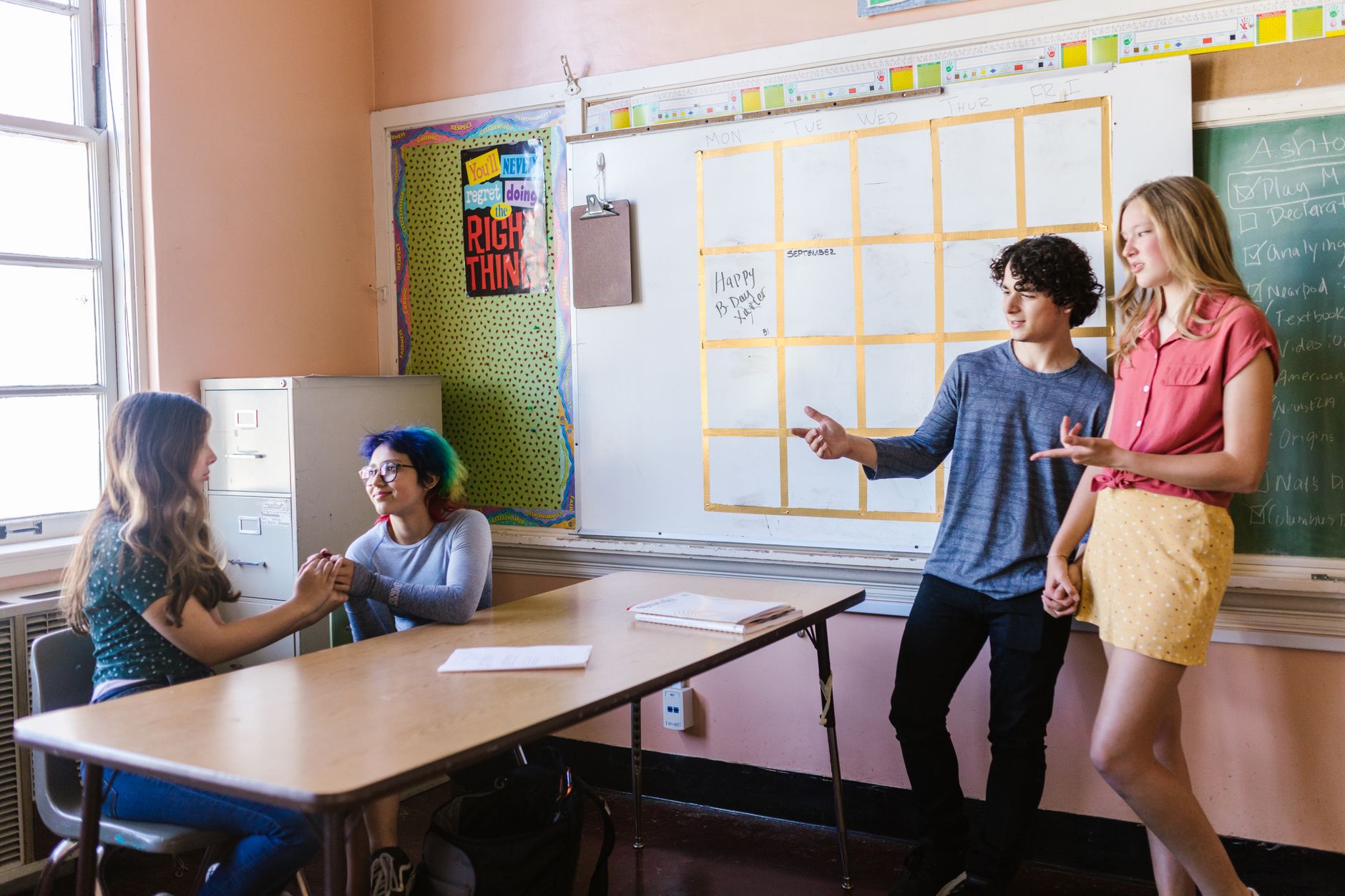 Quatre adolescents discutent dans une salle de classe : deux assises à une table, deux debout devant un tableau blanc.